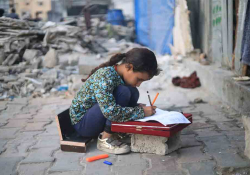 A young girl kneels in the rubble to use a displaced stone as a desk to work on schoolwork