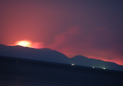 A photograph of a landscape at night, a fire burning ominously behind a hill in the background
