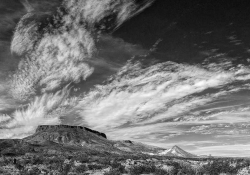 A black and white photograph of a cloud streaked sky above a mesa and the scrublands below