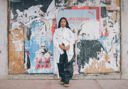 A photograph of a dark skinned woman standing in front of a urban wall