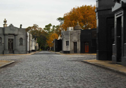 A photograph of a small-stone paved road, lined by ornate mausoleums, inside a tidy cemetary