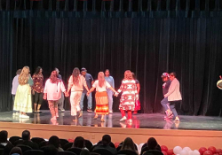 A group of people dance on a bare stage while holding hands