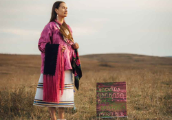 A photograph of a woman in indigenous garb standing on scrub grass hilltop with a painting nearby