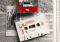 A white cassette with the world Kobold printed on it in red. The cassette lays on a garishly illustrated text