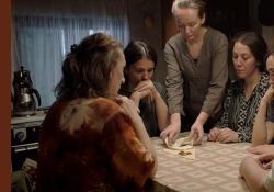A group of women seated around a table. One woman, who is standing, appears to unfolding a cloth to reveal gold within