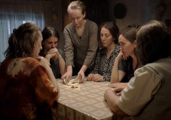 A group of women seated around a table. One woman, who is standing, appears to unfolding a cloth to reveal gold within