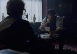 A figure with their back to the viewer looks on as a seated man bemusedly looks at a piece of paper