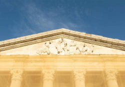 A photograph of a detail near the roofline of an aged marble building. The area has a relief sculpted into including several historical and mythological figures, including Joseph Conrad