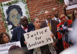 A photograph of a number of African Americans at a press conference. The woman in the center holds a sign that reads, "Justice for Section 14"