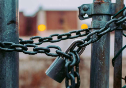 A close-up on a padlock on a chain holding a link fence closed. A building can be made out in the unfocused background