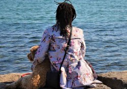 A photograph of a dreadlocked figure sitting with a dog looking out over a body of water