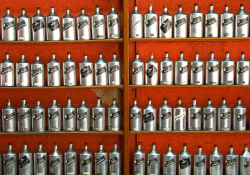 A photograph of long rows of hair tonic in metallic bottles against a red-orange wall