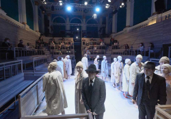 A photograph showing male and female actors dressed in either white trenchcoats or black suits. A socially distanced audience can be seen on bleachers surrounding the stage.