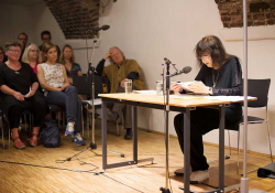 Friederike Mayröcker sits at a table reading with a small group sitting to the side