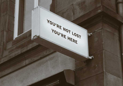 A sign mounted on the corner of a building. The sign reads, ”You are not lost. You are here.”