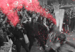 A black and white photo of protesters, mostly female, throwing glitter bombs, which are highlighted in red