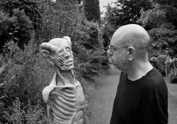 A black and white photograph of Dimitris Lyacos, who is standing on a road running down a wooded lane. He is looking at a grotesque statue that looks like a human with its skin removed.