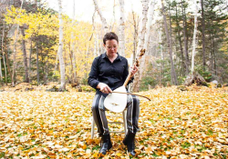 A photograph of Stephanie McKenzie, sitting in an armless chair in a natural setting, while holding a bowed instrument upright in the lap