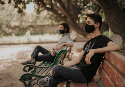 A photograph showing two men, wearing facial masks, sitting on adjacent park benches outside