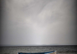 Orange lifejackets piled up on the shore of an island just next to a boat moored nearby. The open ocean and a dark sky looms in the background.