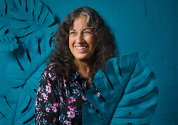 Margarita Engle smiles, holding a broad leaf, painted blue, in front of a blue background