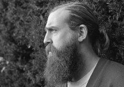 A black and white photograph of a bearded man standing in front of a tall hedge