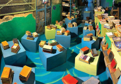 An aerial shot of a warehouse type space, the floor covered with colored boxes with stacks of books on them