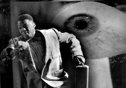 A surreal photograph depicting an African-American man dressed in a suit and carrying a briefcase is running as a giant eye scrutinizes him in the background