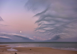 A beach dissolves into a shallow sea, beneath a purpled sky laced with wispy clouds