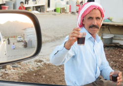 A man wearing a red and white headwrap smiles as he offers the viewer, who is inside of a car, a small, clear glass of tea