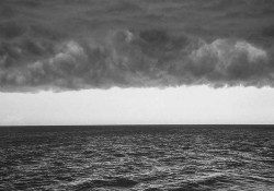 A black and white photo of the sea, dark clouds looming overhead, with the horizon forming the only light between them