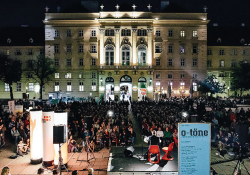 Looking from behind an outdoor stage towards a large crowd assembled to watch a performance with a large building in the background