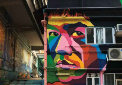 A photograph of a mural on a wall in a crowded urban environment. The mural is of Bruce Lee's face, rendered in bright colors.