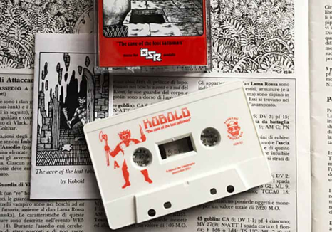 A white cassette with the world Kobold printed on it in red. The cassette lays on a garishly illustrated text