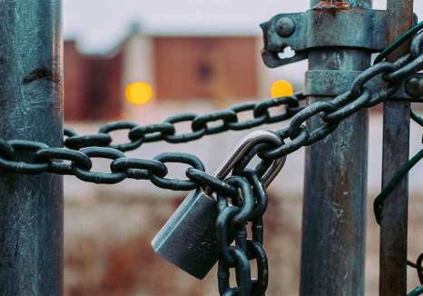 A close-up on a padlock on a chain holding a link fence closed. A building can be made out in the unfocused background