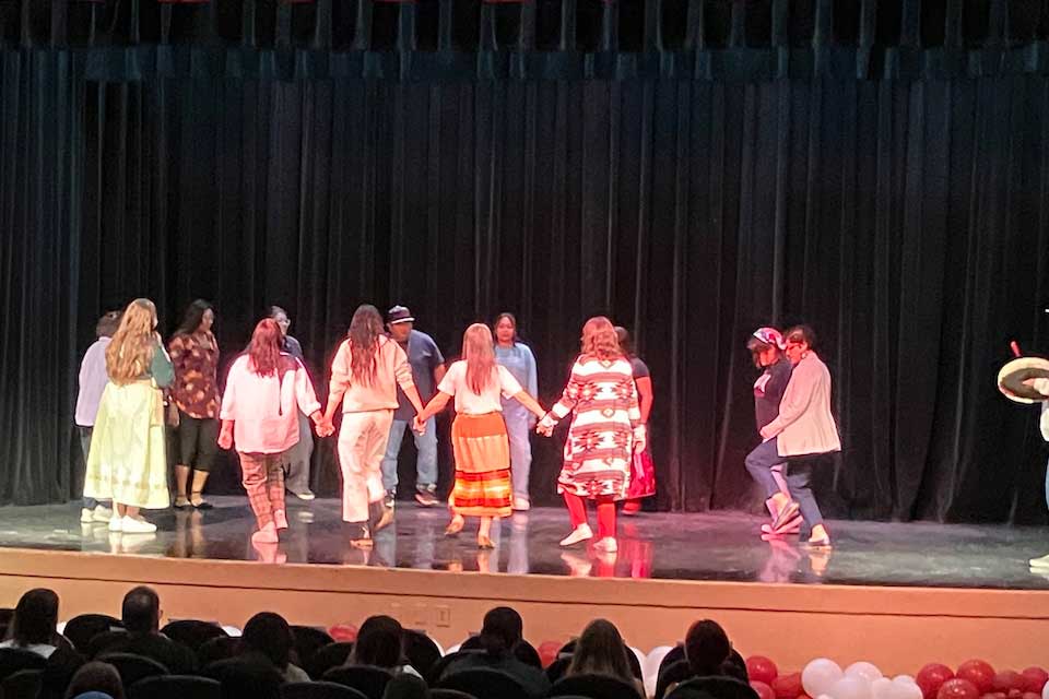 A group of people dance on a bare stage while holding hands